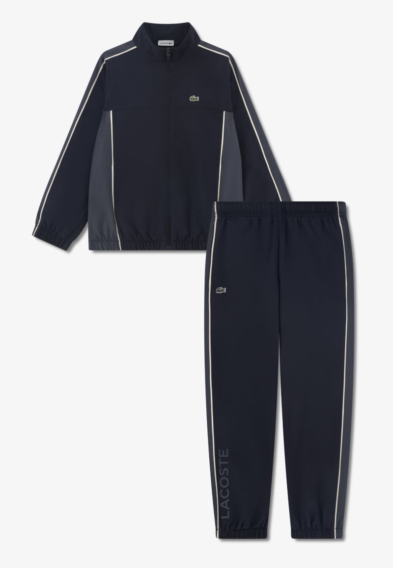 Σετ φόρμας του ναυτικού με ζακέτα με φερμουάρ και λευκή ρίγα, καθώς και παντελόνι που ταιριάζει. Κατασκευασμένο από απαλό ύφασμα, με λεπτομέρεια λογότυπου Lacoste.
