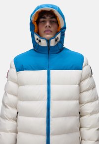 Puffjacka med en blå övre sektion, vit undetermined sektion och orange inre foder. Har en framdragkedja och en huva med dragsko.