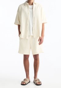 Chemise légère beige clair à manches courtes avec texture, associée à un short assorti, le tout dans une coupe décontractée. Complété par des sandales à lanières marron avec boucles.