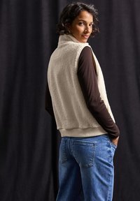 Crèmekleurige fleece vest met hoge kraag, ribbelzoom, gecombineerd met een bruin langetermijn shirt en blauwe jeans, gezien vanaf de achterkant.