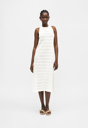 Kvinde iført en hvid ærmeløs strikket midikjole og beige sandaler med sløjfedetaljer på en ensfarvet lys baggrund.