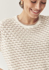 Suéter de punto crema con un patrón de diamantes texturizado, cuello de canalé y mangas cortas. Pendientes de aro dorados visibles en la imagen.