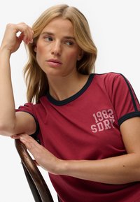 Weinrotes kurzärmeliges T-Shirt mit marineblauem Rand und weißem Aufdruck "1982 SDRY" auf der Brust. Glatte Stoffstruktur, lässige Passform.