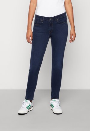 Skinny dark blue jeans with a smooth texture, front pockets, and a button closure, paired with white sneakers featuring green stripes.