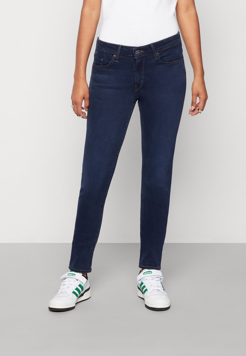 Jean skinny bleu foncé avec une texture lisse, poches avant et fermeture à bouton, associés à des baskets blanches avec des rayures vertes.