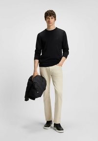Black knit sweater with a round neck, beige chinos, and black sneakers. Holding a black puffer jacket, standing against a light background.