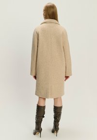 Beige vlněný kabát s širokým límcem, spadlými rameny a rovnou siluetou. Nesený přes vysoké, volné, tmavě hnědé kožené boty.