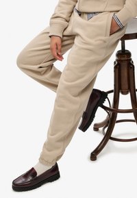 Pantaloni della tuta beige con polsini elasticizzati, dotati di tasche laterali e un piccolo logo ricamato. Abbinati a mocassini bordeaux scuro e calze a coste.