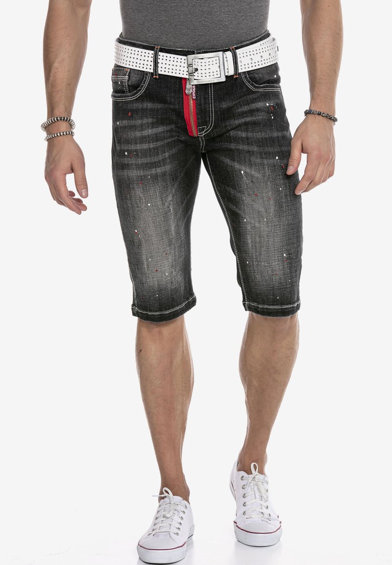 Schwarze Denim-Shorts mit einem ausgewaschenen Finish, rotem Reißverschluss, weißem Nieten-Gürtel und Spritzdetails. Mit umgeschlagenen Säumen und typischem Fünf-Taschen-Design.