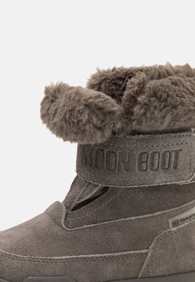 Botte en daim gris avec un col en fausse fourrure, maintenue par une sangle Velcro, possédant une surface texturée et une étiquette spécifique pour la résistance à l'eau.