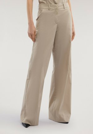 Beige High-Waist-Hose mit weitem Bein, getragen mit schwarzen spitzen Schuhen, gezeigt an einer Person vor einfachem Hintergrund.
