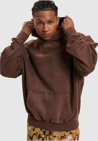 Sudadera marrón de tejido texturizado con un bolsillo canguro y un logo bordado. Combinada con pantalones de patrón de camuflaje.