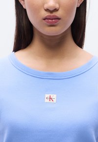 Ανοιχτό μπλε ριγωτό t-shirt με στρογγυλή λαιμόκοψη και ετικέτα με το λογότυπο Calvin Klein που διαθέτει κόκκινες και λευκές λε πτομέρειες στην περιοχή του στήθους.