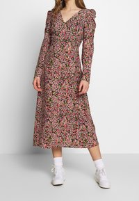 Blommönstrad långärmad klänning i svart med rosa och gröna mönster. Har en V-ringning och plisserade puffärmar, kombinerad med vita sneakers.