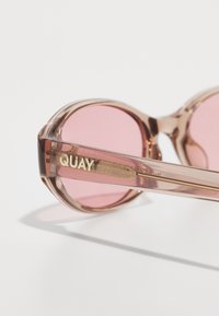 Lunettes de soleil rondes teintées de rose avec une monture translucide de couleur marron clair. Logo "QUAY" doré sur la branche. Texture lisse, design élégant.