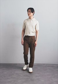 Kortærmet beige polo med lynlås krave, parret med brune snøre bukser og hvide sneakers, stående mod en ensfarvet baggrund.