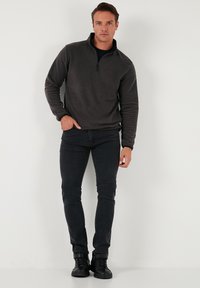 Grå fleecepullover med høj krave og halv lynlås, parret med sorte slim-fit jeans og sorte snørebåndsstøvler, stående på en ensfarvet baggrund.