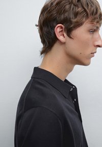 Černá košile s límečkem a knoflíky, která má měkkou texturu a slim fit. Model je zobrazen v profilu na jednobarevném pozadí.