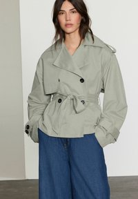 Trench coat grigio chiaro, doppiopetto, con cintura in vita, grandi bottoni neri e ampie maniche, abbinato a pantaloni in denim blu larghi.