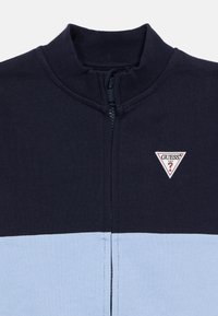 Μπλε ναυτικό και ανοιχτό μπλε φούτερ με φερμουάρ και ριμπ γιακά, με τριγωνι κό λεκτικό λογότυπο "GUESS?" στη αριστερή πλευρά. Απαλή υφή.