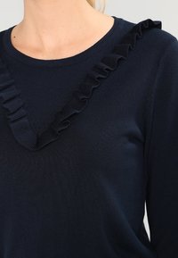 Marinblå tröja med V-ringning och texturerad volangdetalj längs halslinjen. Mjuk tyg med en slät yta och figursydd design.