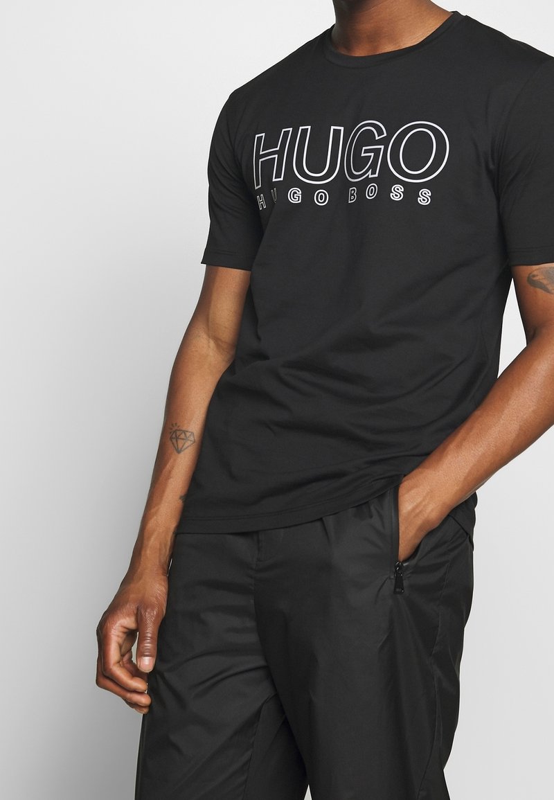 Musta puuvillapaita, jossa on rohkea valkoinen graafinen logo "HUGO" rinnassa. Lyhyet hihat ja klassinen pyöreä kaula-aukko.