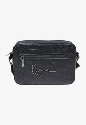 Black crossbody bag in textured synthetic material. Features a front zip pocket and silver-tone logo accent. Rectangular shape with adjustable strap.