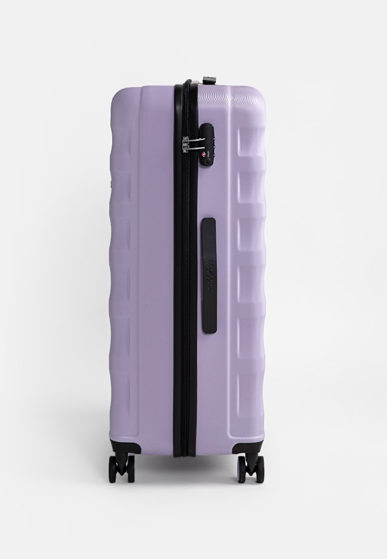 Valise rigide en lavande clair, avec une surface texturée, une fermeture éclair noire, une poignée latérale et quatre roues pour faciliter le déplacement.