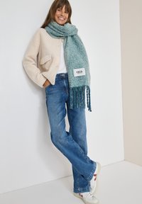 Beige fleecejakke med en frontlomme, blå franstørret tørklæde, blå jeans og hvide sneakers med røde detaljer, sat mod en neutral baggrund.