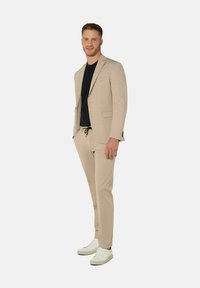 Beige Zweiteiler-Anzug mit schlanker Passform, strukturierter Stoff, Ein-Knopf-Blazer und Hose mit Kordelzug, kombiniert mit weißen Sneakers.