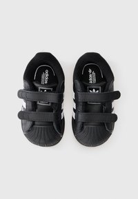 Des chaussures pour tout-petits en cuir noir avec deux bandes Velcro, un embout texturé et trois rayures blanches sur les côtés.