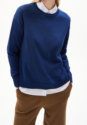 Personne portant un pull bleu à col rond par-dessus une chemise blanche boutonnée, avec un pantalon marron, les mains dans les poches, sur un fond blanc uni.