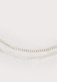 Tre collane sovrapposte: una catena d'oro con pietre preziose trasparenti, un filo di perle bianche rotonde e una delicata catena d'oro con piccoli cristalli trasparenti.