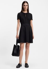 Μαύρο φόρεμα μέχρι το γόνατο με κοντά μανίκια, με λαιμόκοψη σε σχήμα κλειδιού. Συνδυάζεται με μαύρη δερμάτινη τσάντα και επίπεδα παπούτσια. Λείο ύφασμα.