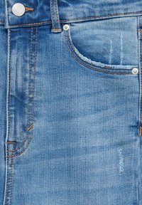 Gros plan sur un jean en denim bleu montrant la poche avant, la boucle de ceinture, des rivets métalliques et un bouton métallique brandé avec des détails de surpiqûres orange.
