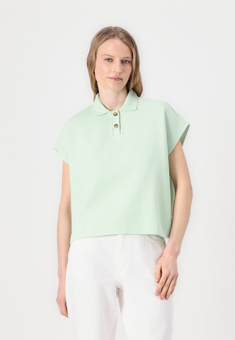 Lichtgroene polo met korte mouwen, ontspannen pasvorm en knoopsluiting met twee knopen. Gemaakt van zacht materiaal. G gedragen met witte broek.
