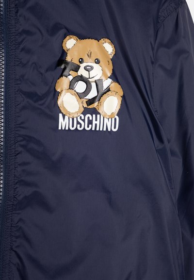 Jūros mėlynos spalvos vėjui atsparus paltas su tekstūruotu meškiuko pleistru, ant kurio parašyta "TOY", o po meškiuku baltuoju šriftu esantis "MOSCHINO" logotipas.