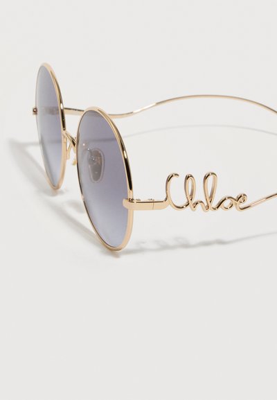Στρογγυλά γυαλιά ηλίου με χρυσές μεταλλικές κορνίζες και γκρι φακούς με απόχρωση, με την υπογραφή "Chloe" στα μπράτσα.