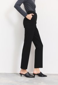 Zwarte, op maat gemaakte broek met rechte pijpen, zijzakken en een hoge taille, gedragen met zwarte mules met open teen, tegen een effen achtergrond.