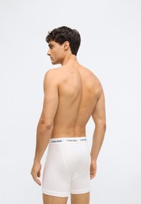Muž standing bez trička, oblečený v bílých boxerkách Calvin Klein, stojící vlevo s viditelnými svaly zad a logem v pase na jednobarevném pozadí.