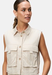 Beige bezrukávová košile na knoflíčky s dvěma kapsami na hrudi, vyrobená z lehkého materiálu. Má klasický límec a jemnou texturu.