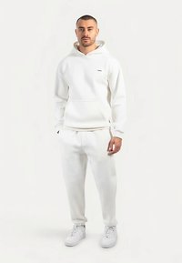Weißer Hoodie und passende Sweatpants aus weichem Stoff. Der Hoodie verfügt über eine Kängurutasche und ein kleines Logo. Das Modell trägt weiße Sneaker.
