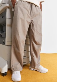 Pantaloni loose-fit di un beige chiaro, realizzati in un tessuto morbido, con tasche laterali e una cintura elastica, abbinati a scarpe da ginnastica bianche su un pavimento giallo.