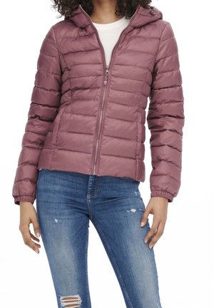 Woman wearing a mauve quilted zip-up jacket with a hood and blue distressed jeans.