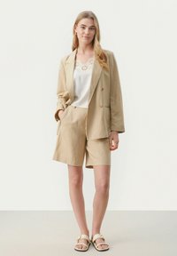 Beige skræddersyet blazer med matchende shorts, lavet af let stof. Toppen har en blondekant og er parret med cremefarvede sandaler.