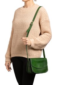 Borsa a tracolla verde in pelle con chiusura a pattina, forma strutturata, tracolla regolabile e texture liscia. Indossata sopra un maglione chiaro.