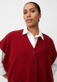 Gilet rosso a maglia con scollo a V, maniche corte e bottoni neri, indossato sopra una camicia bianca con colletto, caratterizzato da una texture morbida.