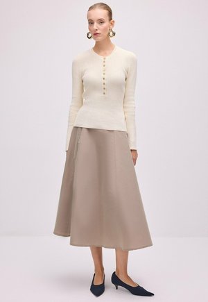 Femme portant un pull crème à manches longues boutonné, une jupe midi beige en ligne A, de grandes boucles d'oreilles anneaux dorées et des chaussures noires à talons bas.