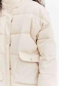 Nafouklá bílá bunda s texturovaným povrchem, vybavená zapínáním na knoflíky, postranní kapsou a stojatým límcem.