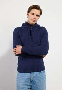 Marinblå stickad huvtröja med hög krage, dragsko och ribbade ärmslut. Kombineras med ljusblå denimjeans. Strukturerad yta genom hela plagget.